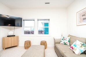 A living room with a flat-screen TV, two windows, a sofa with decorative pillows, a wooden cabinet, and two wicker stools. - The Salty Beagle - 4BR Home with Pool; Ocean & Sound Views (Surf City)