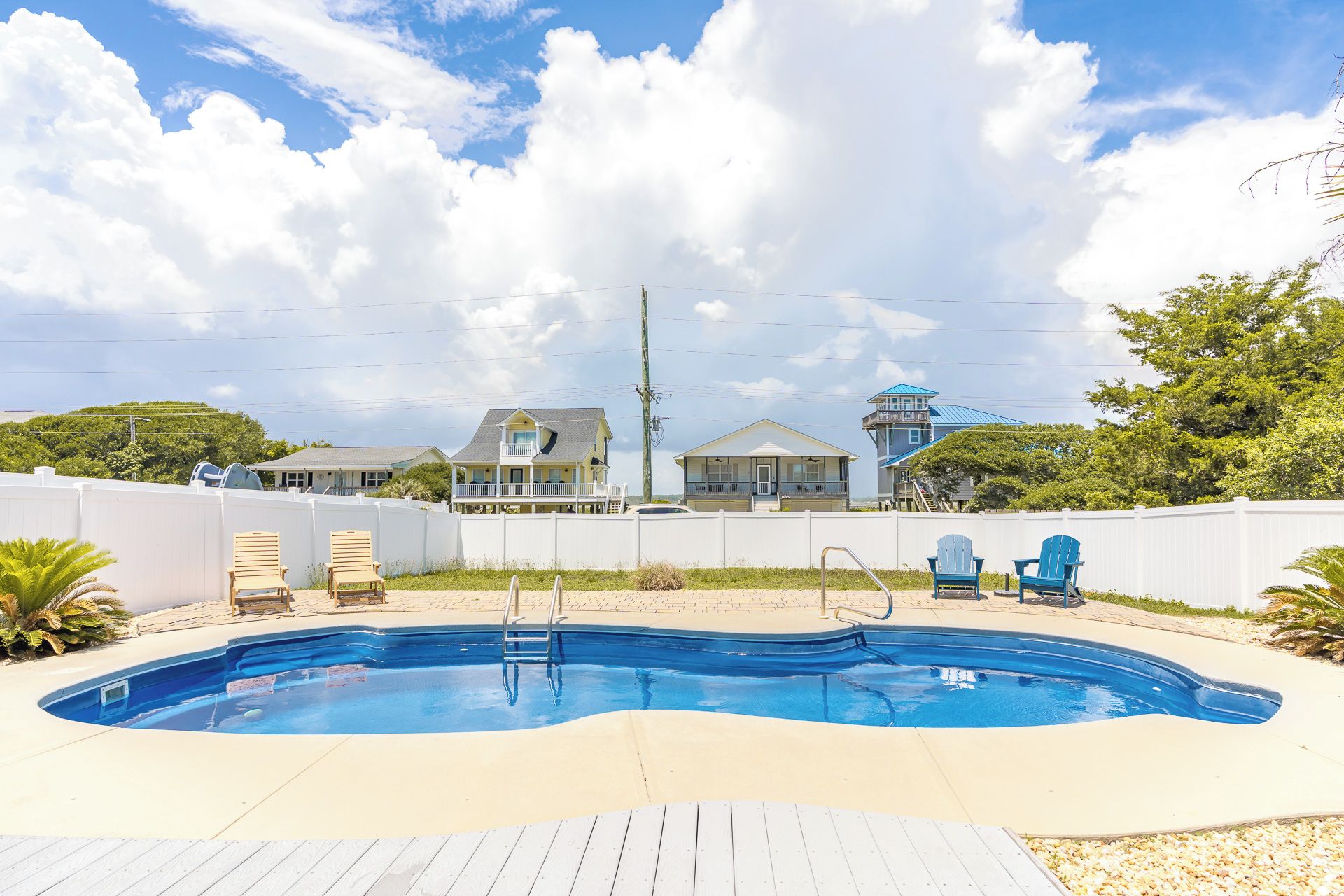 A swimming pool with a wooden deck, surrounded by a white fence, with houses in the background.