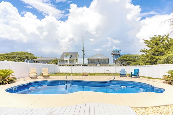 A swimming pool with a wooden deck, surrounded by a white fence, with houses in the background.