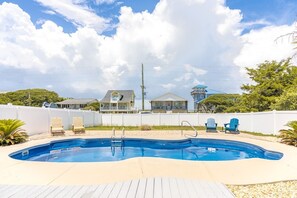 A swimming pool with a wooden deck, two blue Adirondack chairs, and a white fence.