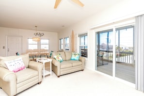 A spacious living room with a large sliding glass door leading to a balcony, a dining area with wooden chairs, and a ceiling fan. - The Salty Beagle - 4BR Home with Pool; Ocean & Sound Views (Surf City)