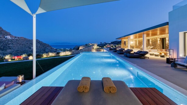 A infinity pool with a view of the sea and a stone building with outdoor seating. - Remarkable 7-bed Villa in Mikonos - Villa Ataraxia (Mykonos)
