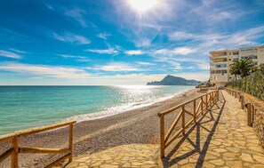 Une promenade en bord de mer avec une plage de galets, une rambarde en bois et une vue dégagée sur la mer et les montagnes lointaines.