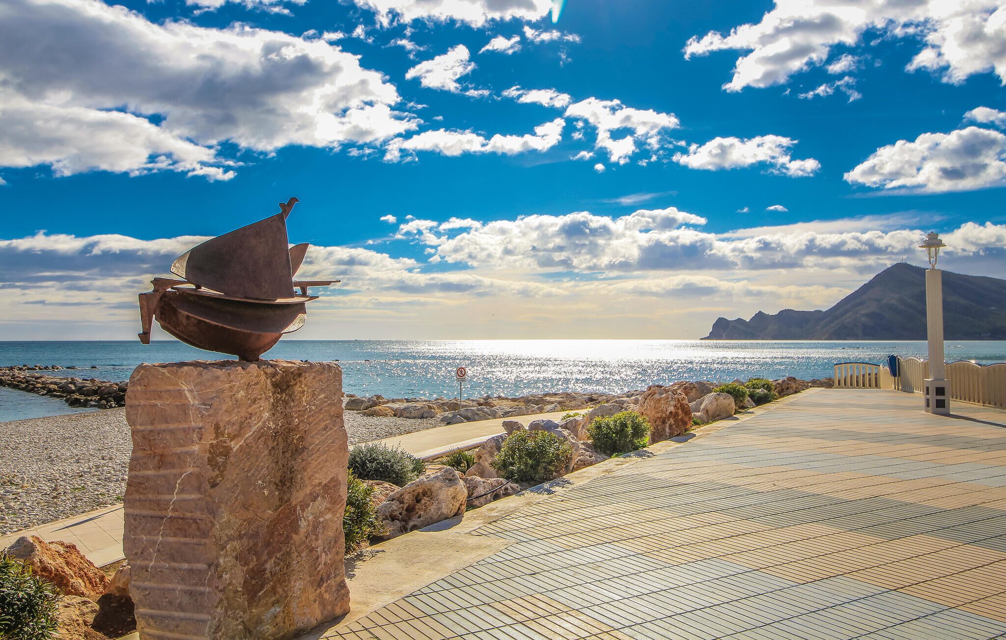 A coastal walkway with a metal sculpture, rocky terrain, and a clear view of the sea and distant mountains.