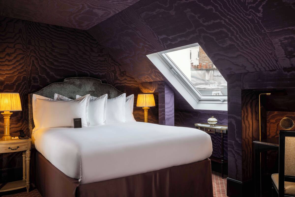 Quarto de hotel com uma cama grande, duas luminárias de cabeceira, uma mesinha com uma lâmpada e um claraboia.
