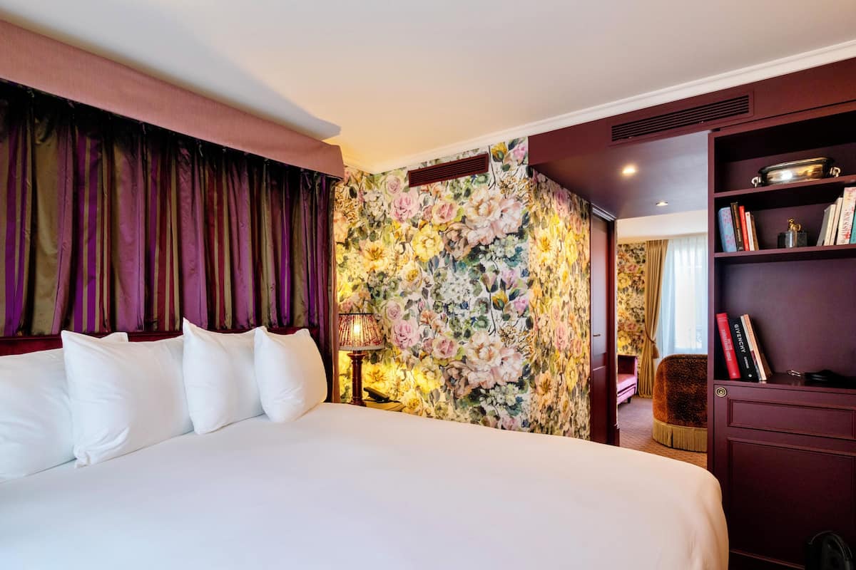 Quarto de hotel com cama grande, papel de parede floral, estante e janela com cortinas.