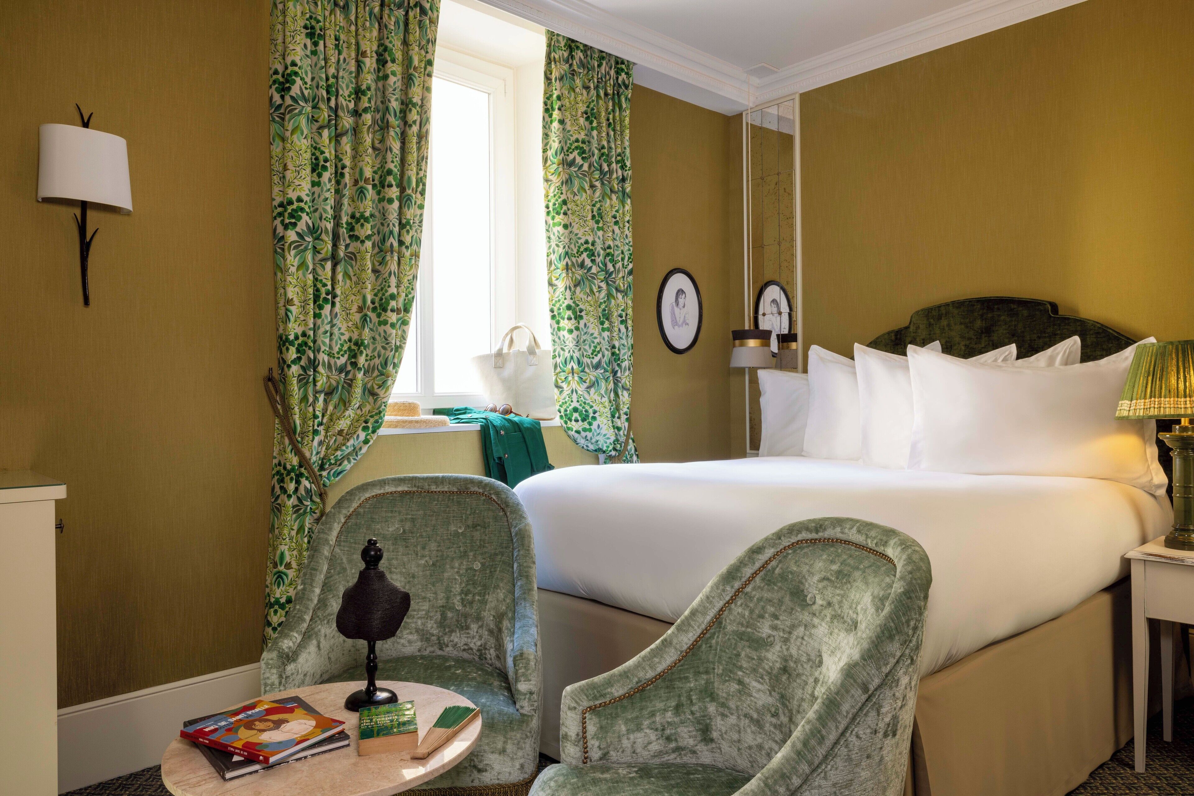 A hotel room with a bed, two chairs, a small table with books, a lamp, and curtains.