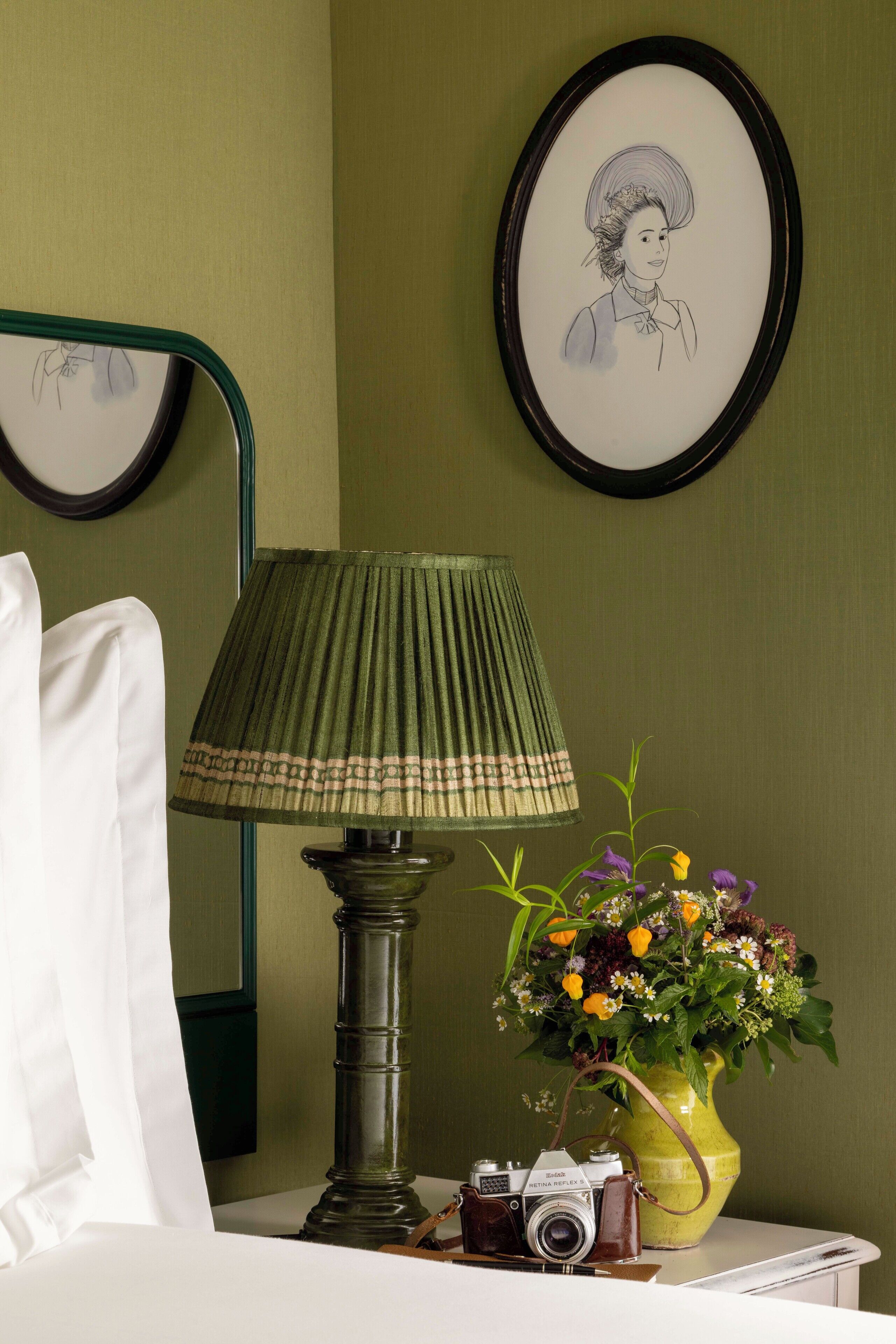 A neatly made bed with white linens, a green lamp, a vintage camera, and a vase of flowers on a bedside table.
