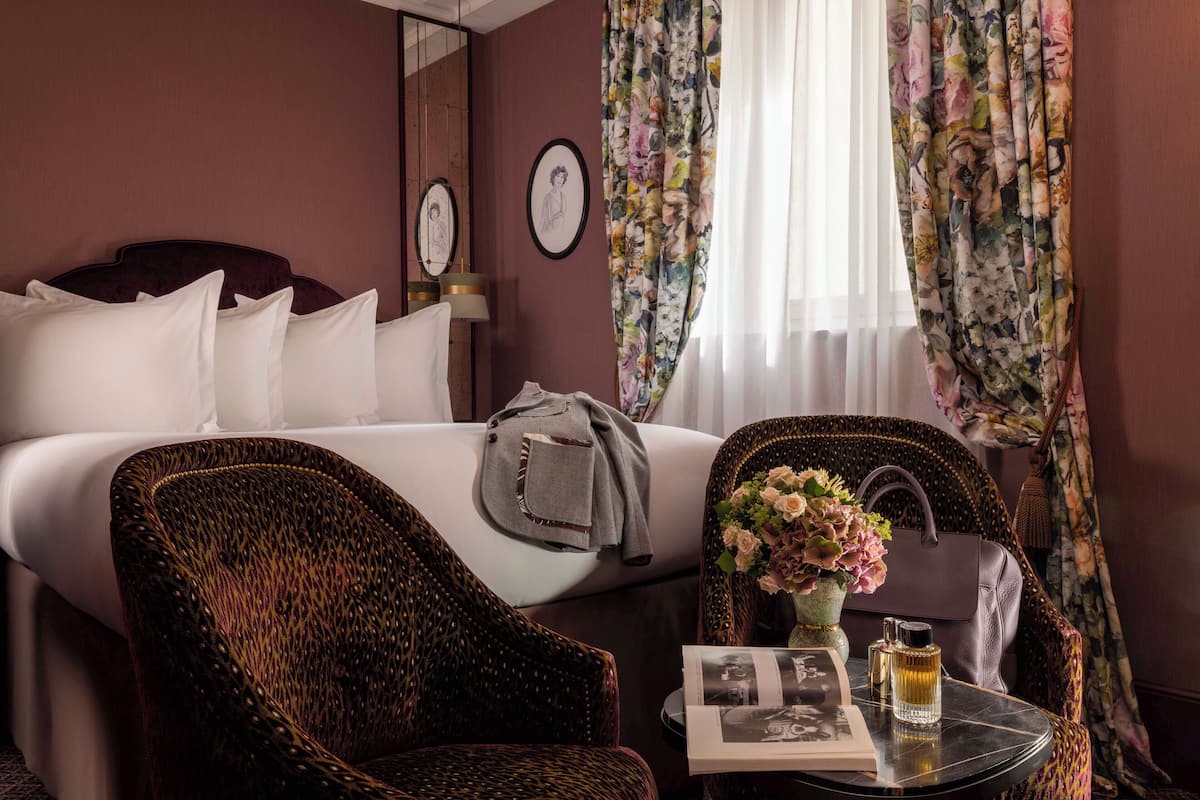 Quarto de hotel com cama, poltrona, mesa com um vaso e perfume, e cortinas.