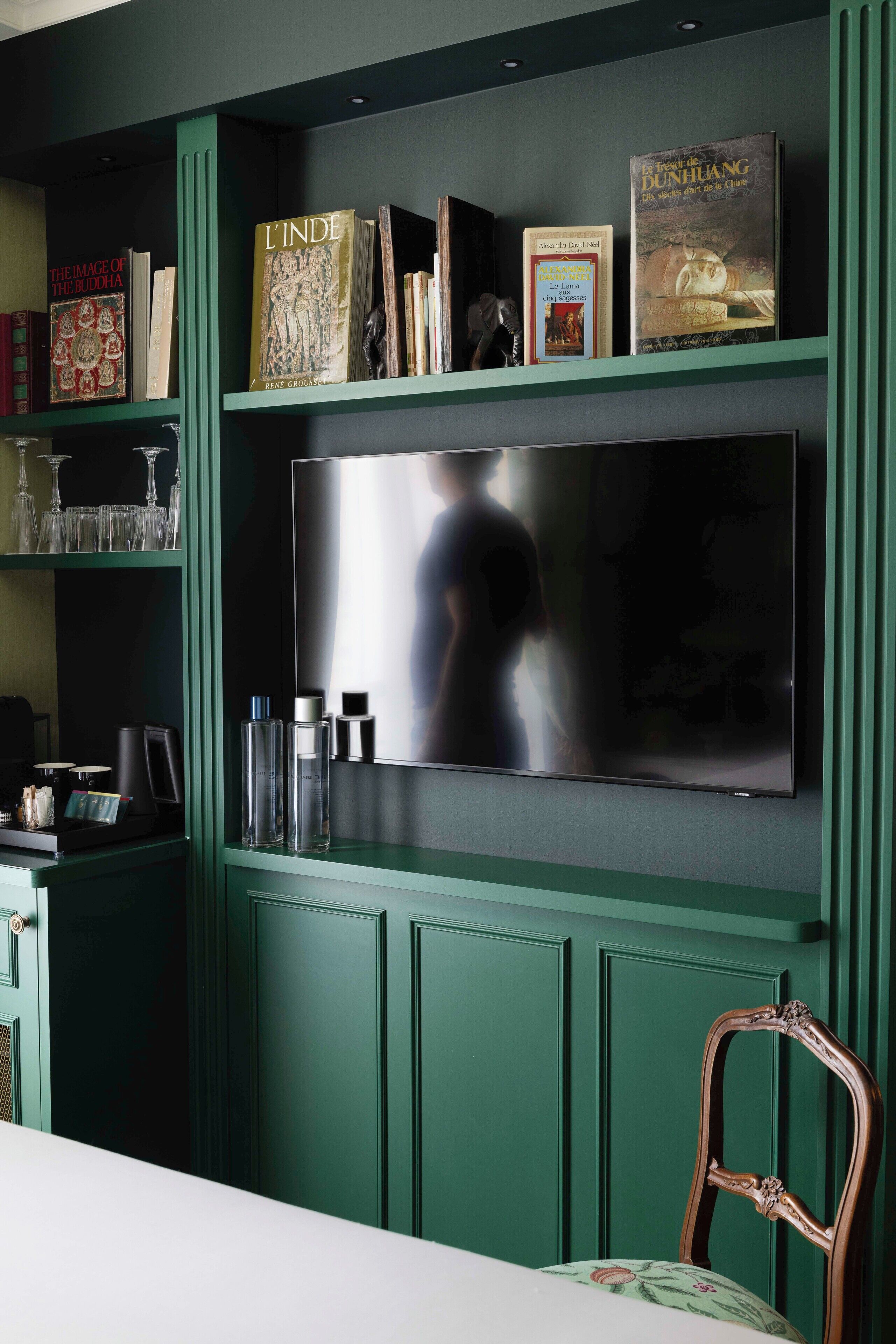 Une pièce avec un grand téléviseur à écran plat fixé sur un mur vert, une chaise en bois et une étagère contenant des livres et des objets décoratifs.