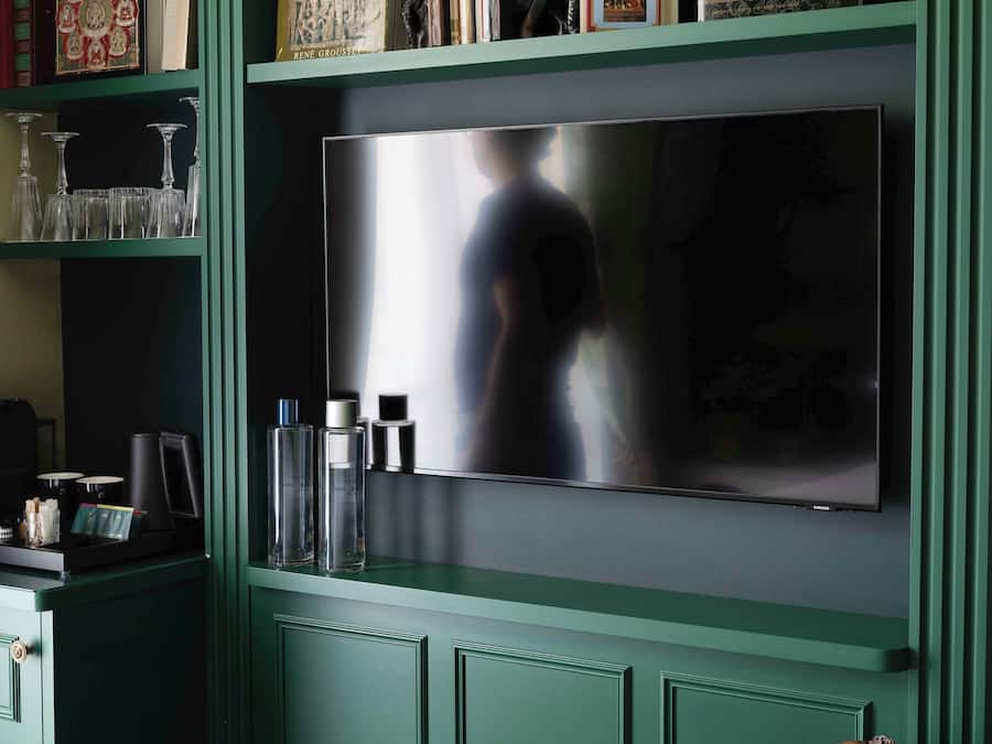 A room with a large flat-screen TV mounted on a green wall, a wooden chair, and a shelf with books and decorative items.
