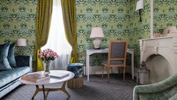A room with a patterned wallpaper, a green sofa, a round coffee table with a book, a small side table with a lamp, a chair, and a fireplace.