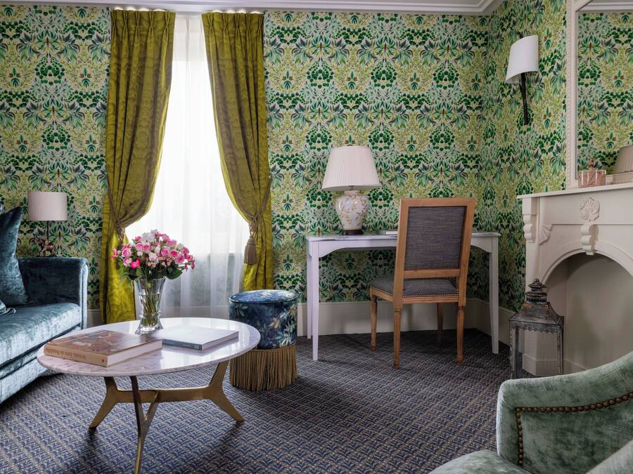A room with a patterned wallpaper, a green sofa, a round coffee table with a book, a small side table with a lamp, a chair, and a fireplace.