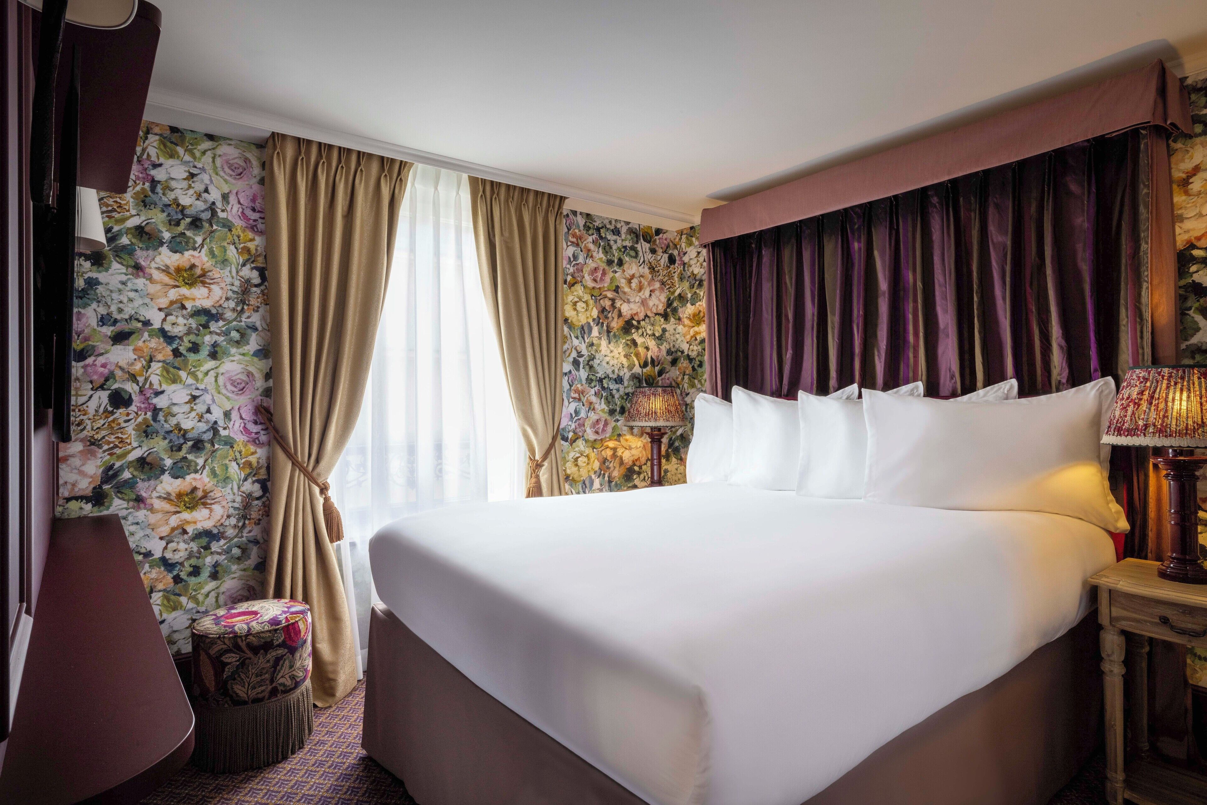 Une chambre d’hôtel avec un grand lit, un petit tabouret, une lampe et un papier peint à motifs floraux.