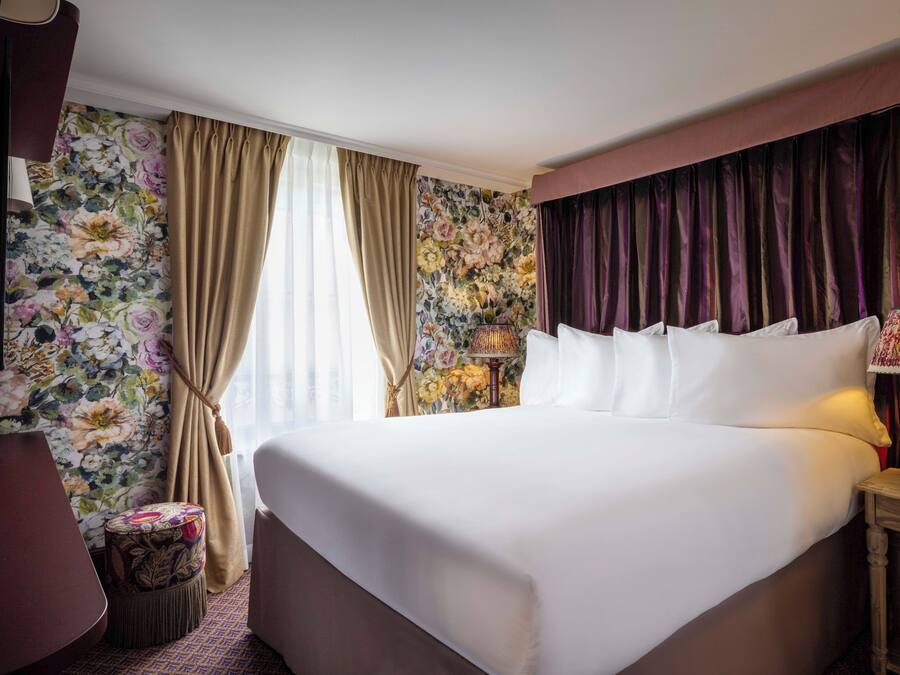 Une chambre d’hôtel avec un grand lit, un petit tabouret, une lampe et un papier peint à motifs floraux.