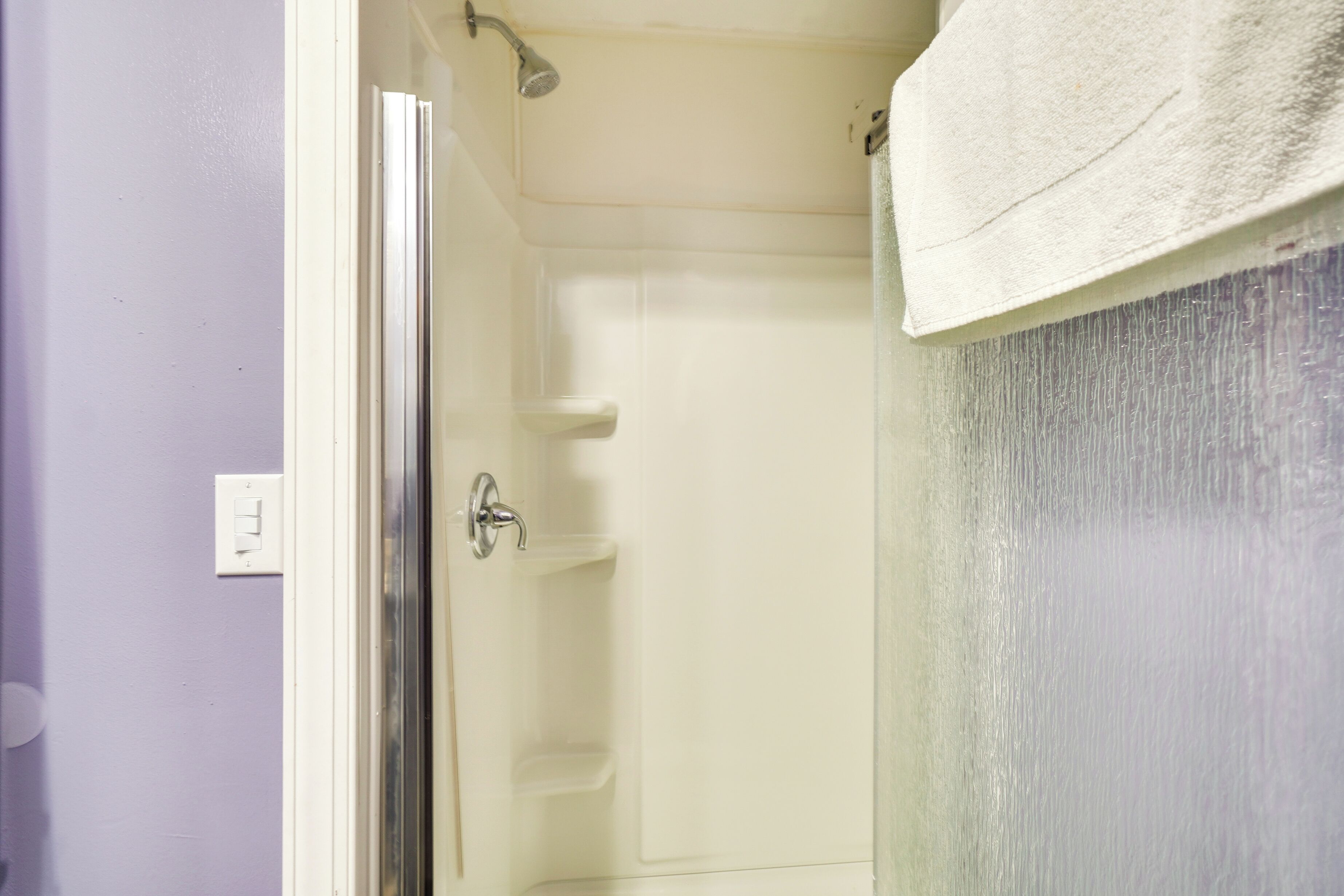 Une douche avec une porte en verre, une serviette accrochée au mur et un mur blanc avec un interrupteur.