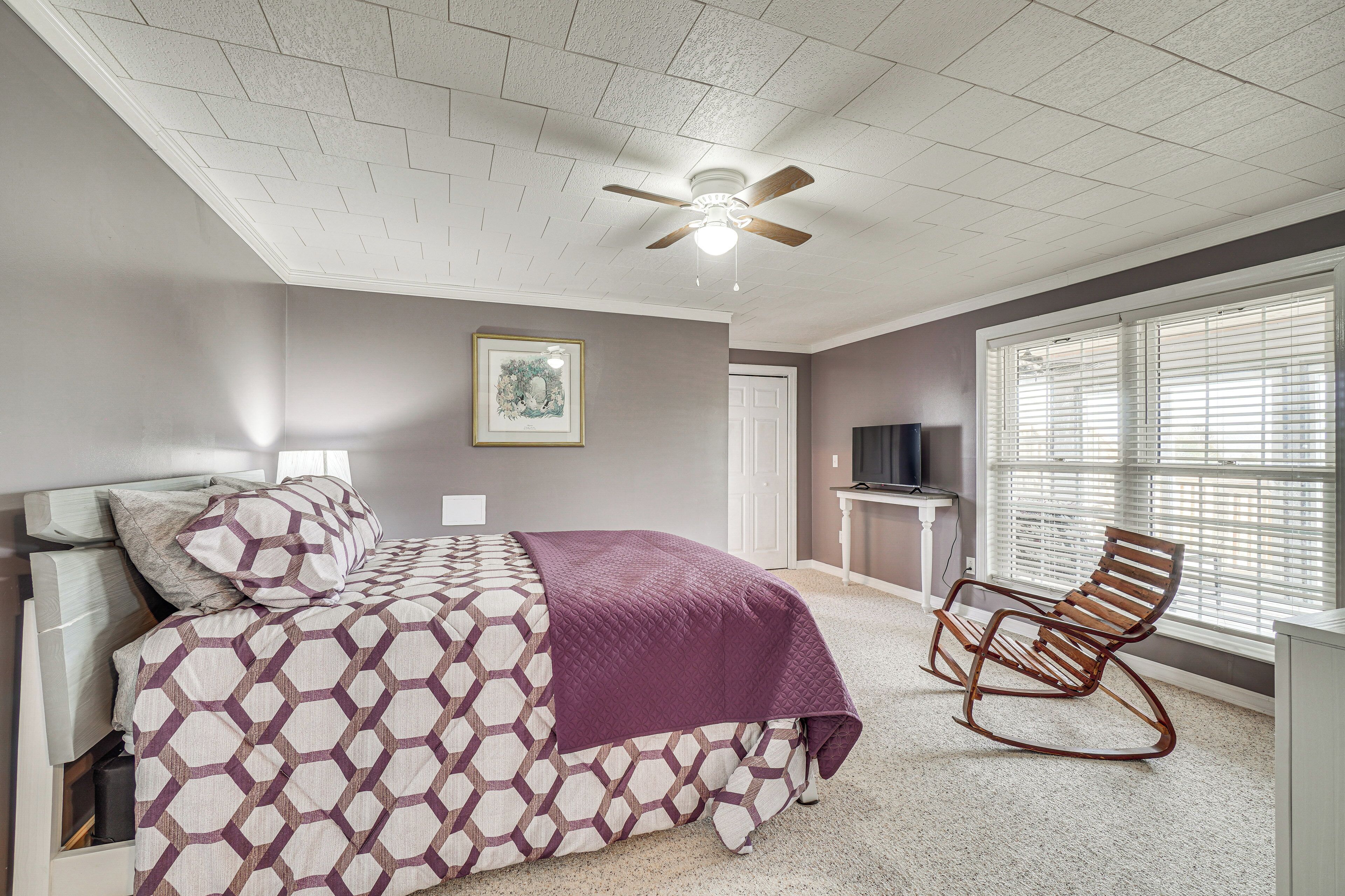 A bedroom with a bed, a rocking chair, a ceiling fan, a window with blinds, and a framed picture on the wall.