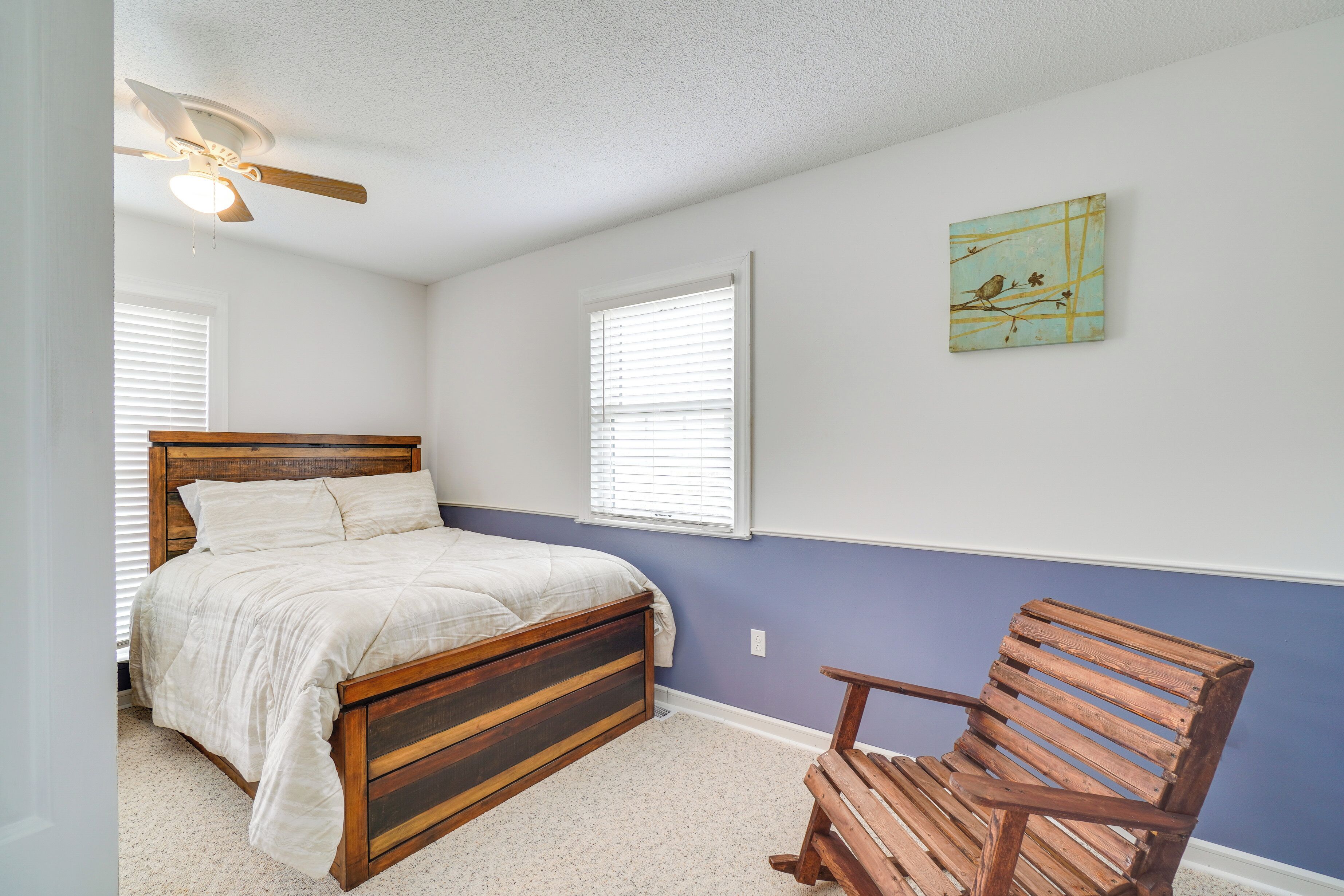 Une chambre à coucher avec un lit en bois, une chaise en bois, un ventilateur de plafond et un tableau accroché au mur.