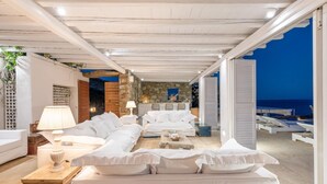 A modern, well-lit room with a large white sofa, a small table, and a lamp. There is a stone wall and a view of the sea. - Inviting 8-bed Villa in Ermoupoli - Villa Blueros (Syros)
