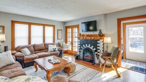 A spacious living room with a fireplace, a television mounted on the wall, and a large window with blinds.
