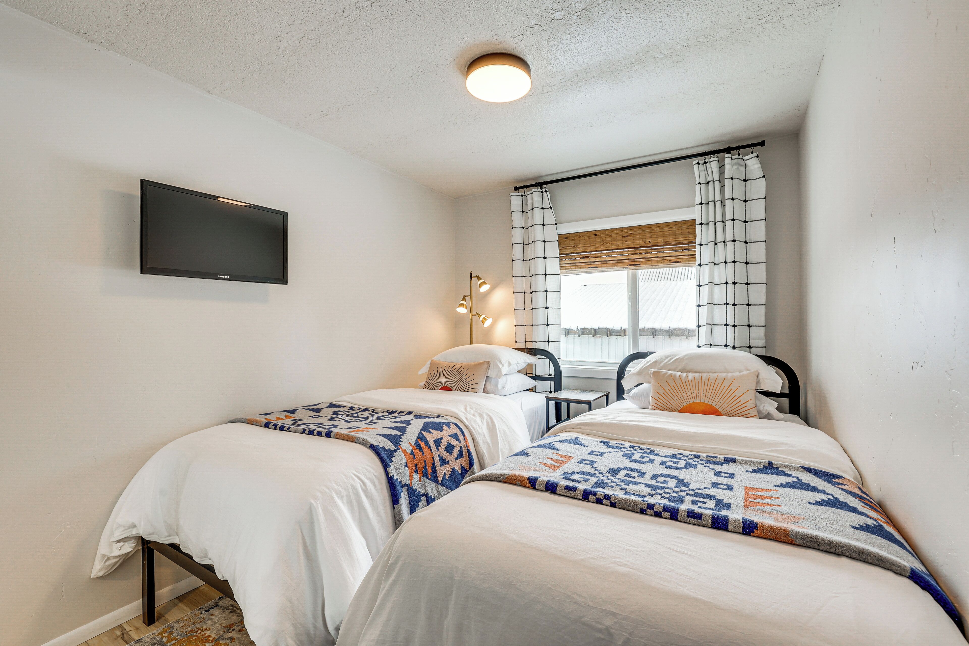 A bedroom with two beds, a flat-screen TV mounted on the wall, a window with curtains, and a small table with a lamp.
