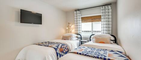 A bedroom with two beds, a flat-screen TV mounted on the wall, a window with curtains, and a small table with a lamp.