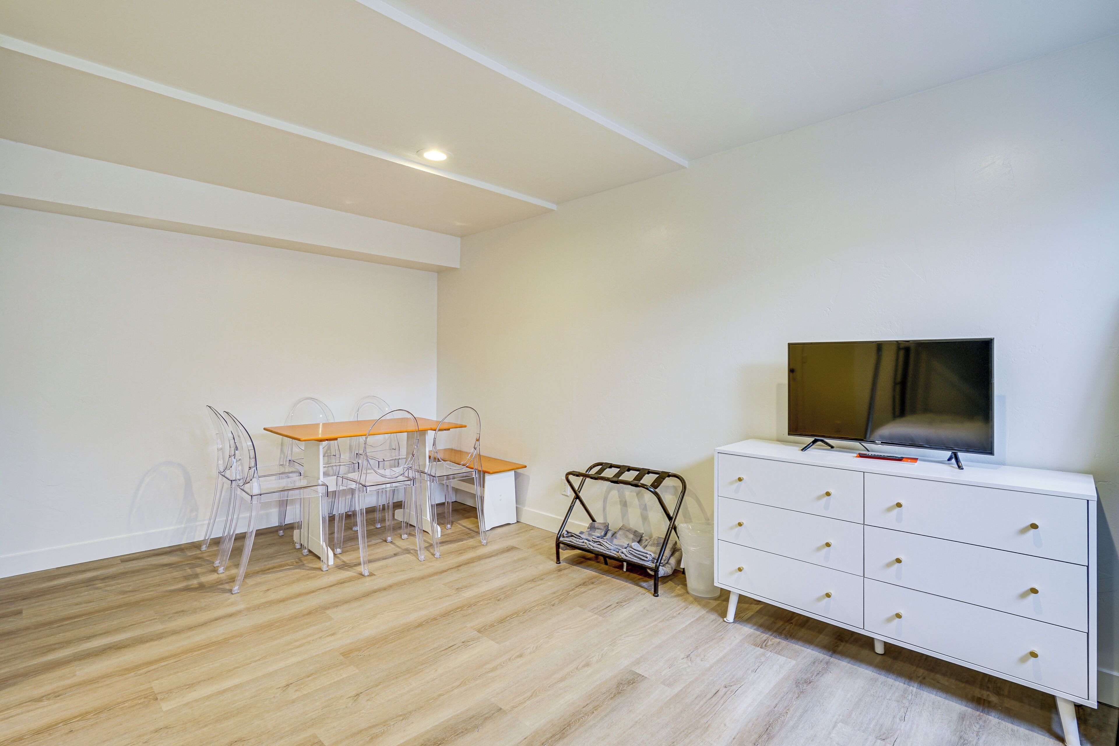 Una habitación con piso de madera, un aparador blanco con cajones, un televisor encima y una mesa con sillas.