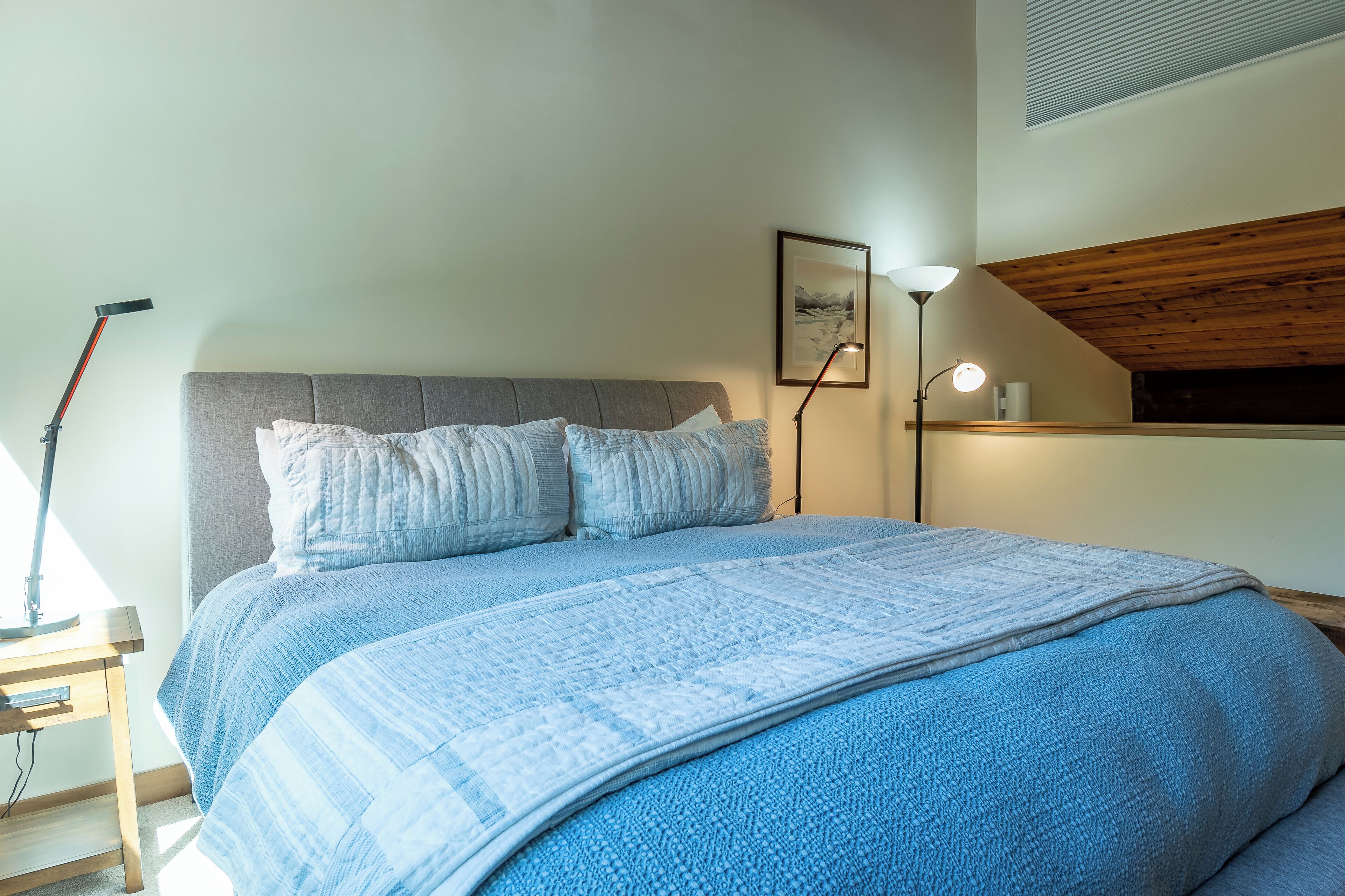 A bedroom with a large bed, a bedside lamp, a picture on the wall, and a wooden ceiling.