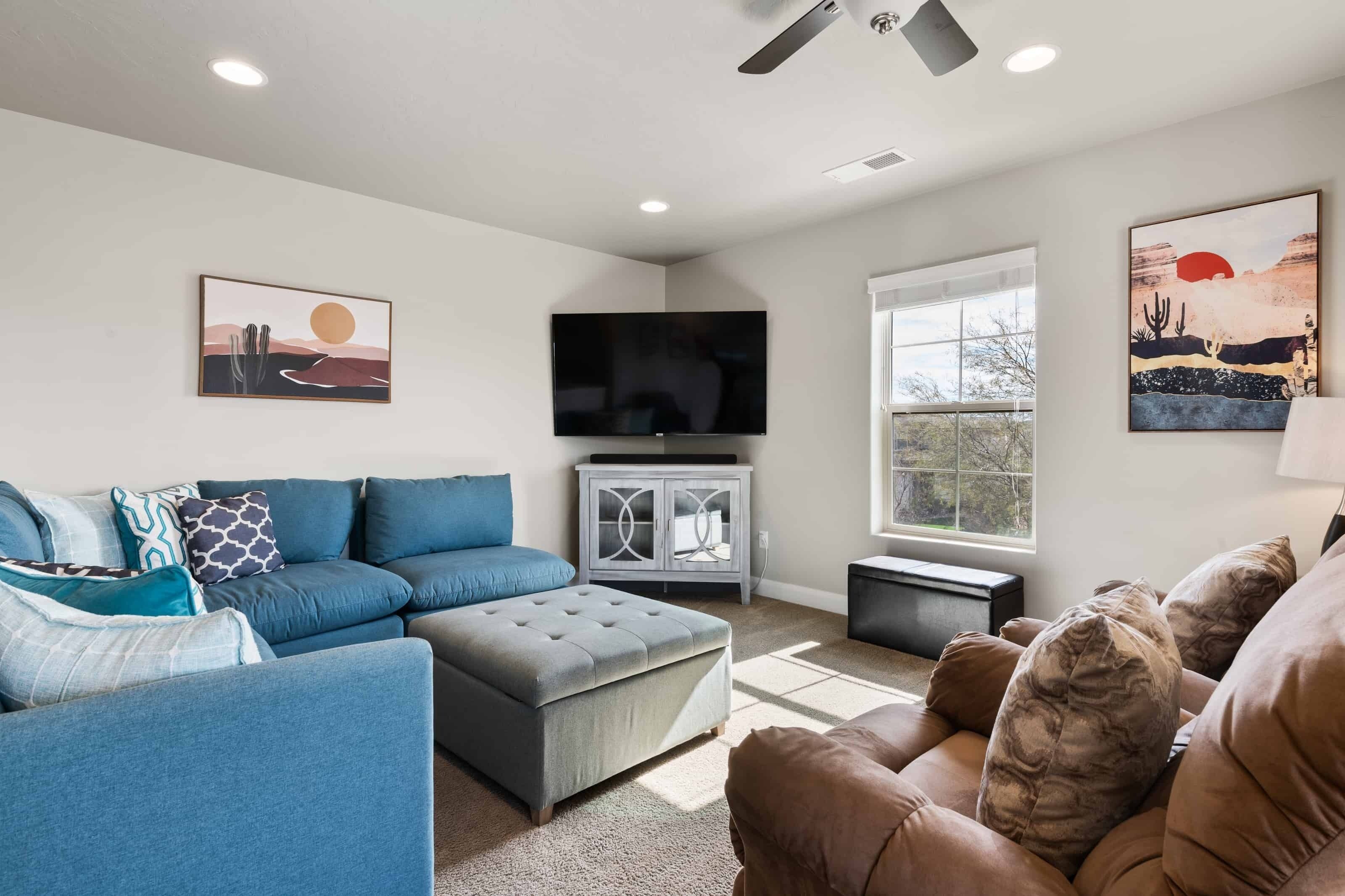 A living room with a blue sofa, a grey ottoman, a television, and artwork on the walls.