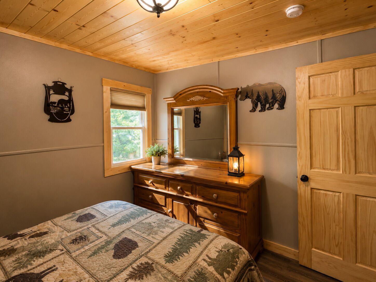 A bedroom with a wooden dresser, a bed with a floral quilt, a window with blinds, a mirror, and a door.