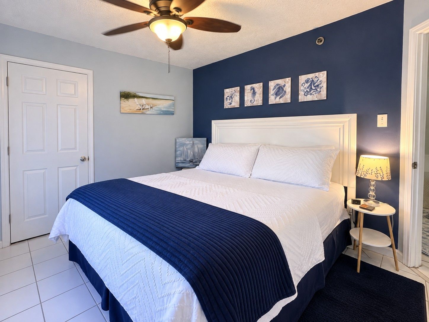 A bedroom with a bed, a ceiling fan, a nightstand with a lamp, and artwork on the wall.