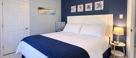 Un dormitorio con cama, ventilador de techo, mesita de noche con lámpara y una obra de arte en la pared.