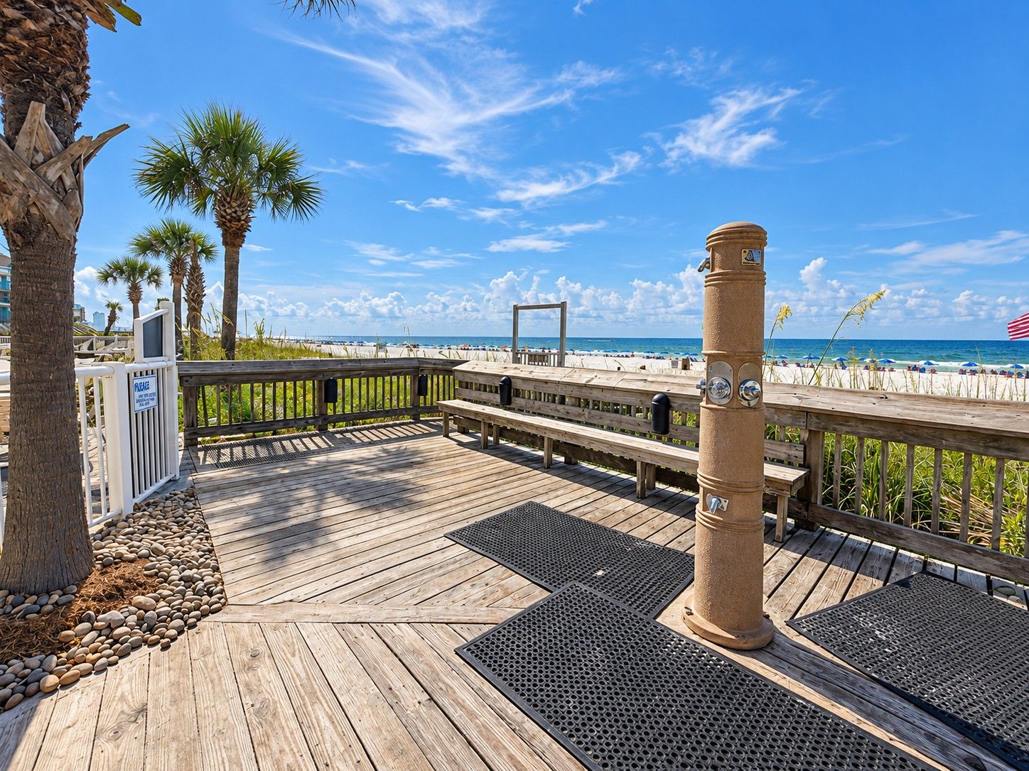 A wooden deck with a railing overlooking a beach, palm trees, and a clear sky.