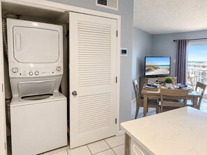 A compact room with a washing machine, a closed door, a dining area with a table and chairs, and a television displaying a beach scene.