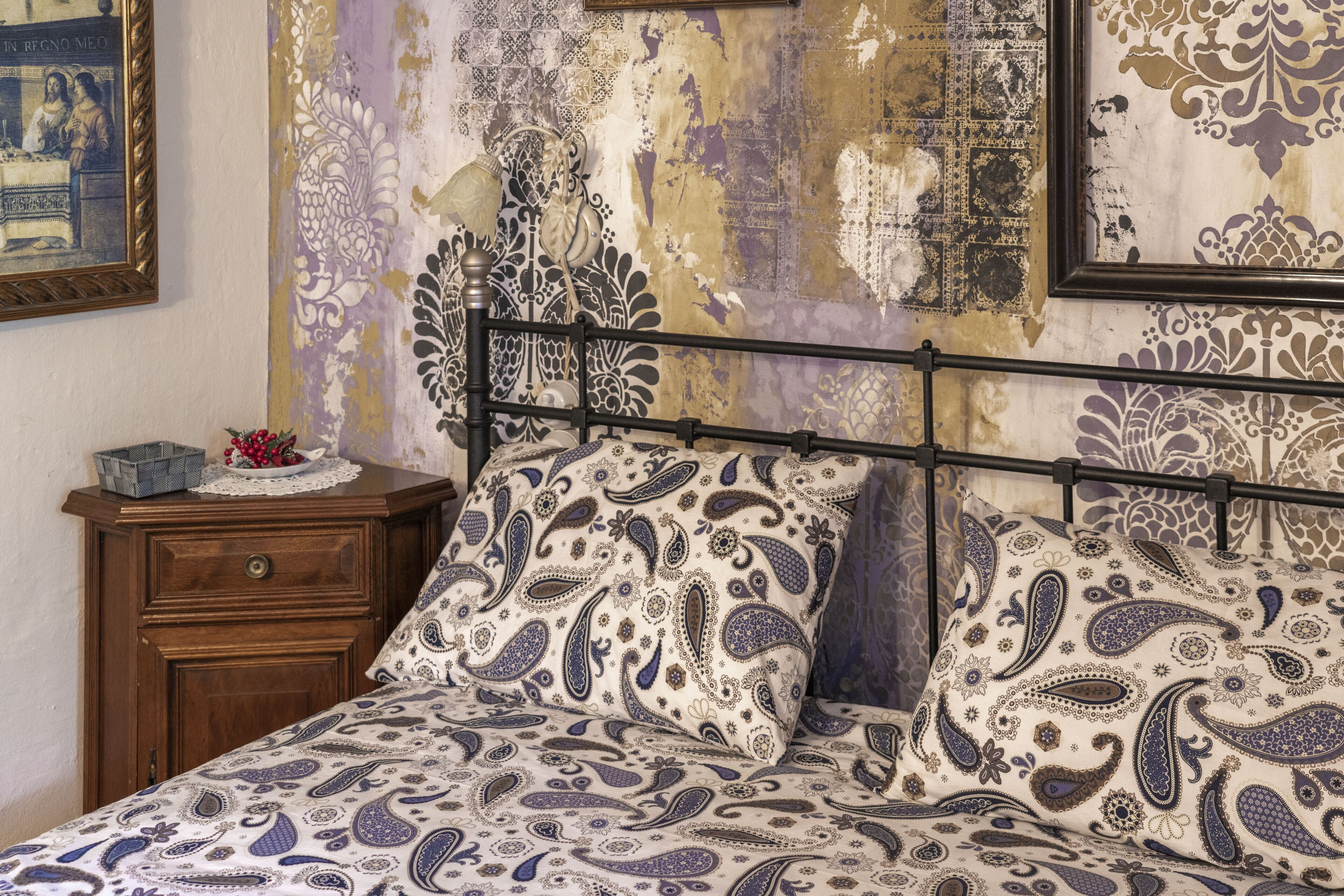 A bed with a patterned bedspread, a wooden nightstand with a basket and a small table, and a wall with a decorative tapestry and a framed picture.
