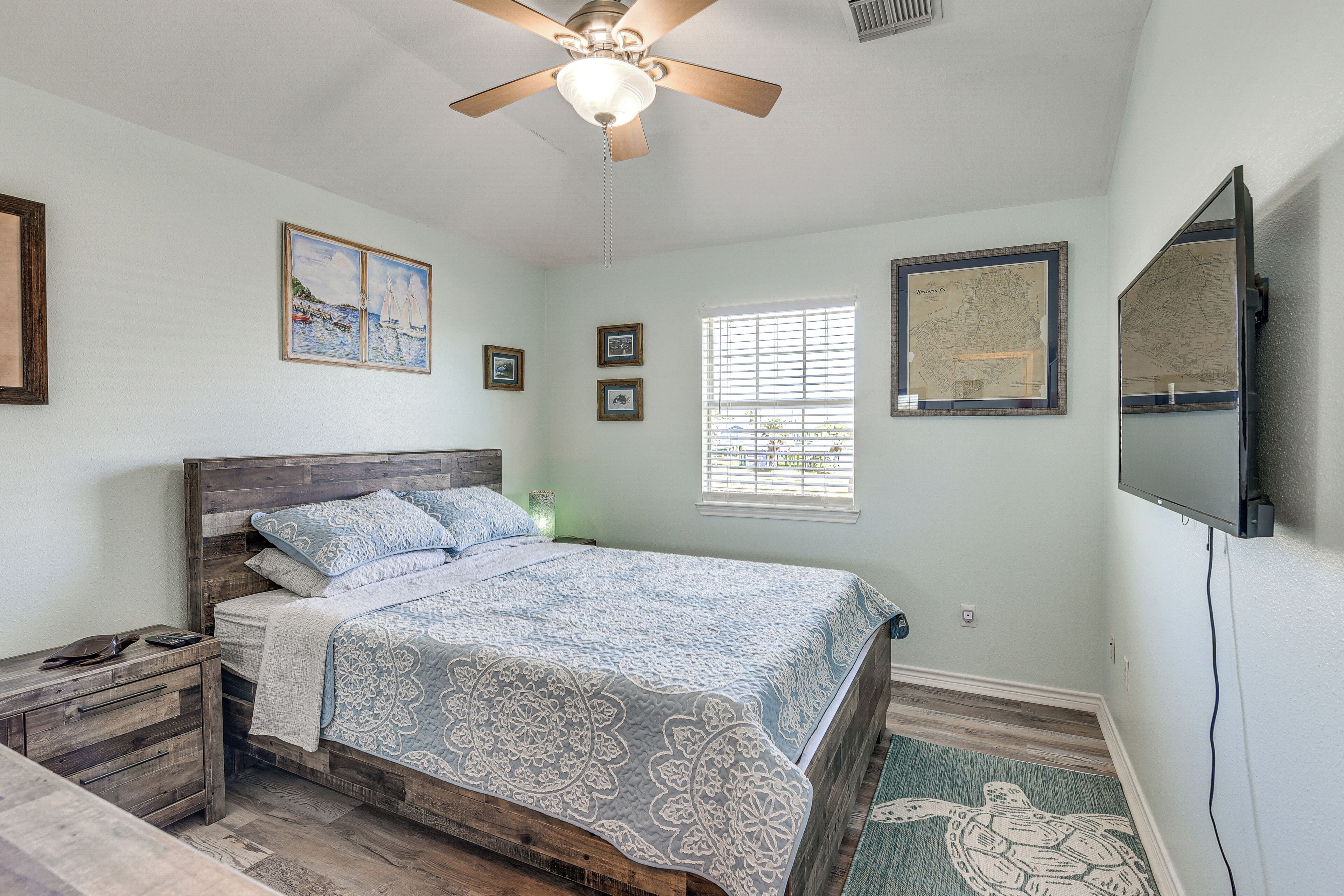 A bedroom with a wooden bed, a ceiling fan, framed artwork, a window with blinds, and a television mounted on the wall.