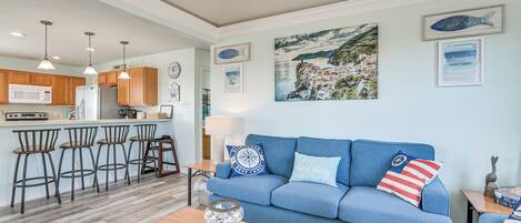 Una cocina moderna con armarios de madera, una zona de bar con taburetes, un sofá azul con almohadones de temática náutica y un cuadro grande de motivos costeros en la pared.
