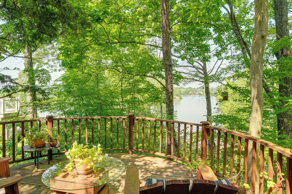 A wooden deck with a glass table and chairs, surrounded by a wooden railing, overlooking a lake.