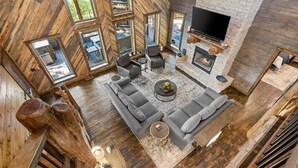 Un vaste salon aménagé dans un style industriel, avec un grand canapé d’angle gris, une cheminée et un téléviseur à écran plat fixé à un mur en briques.