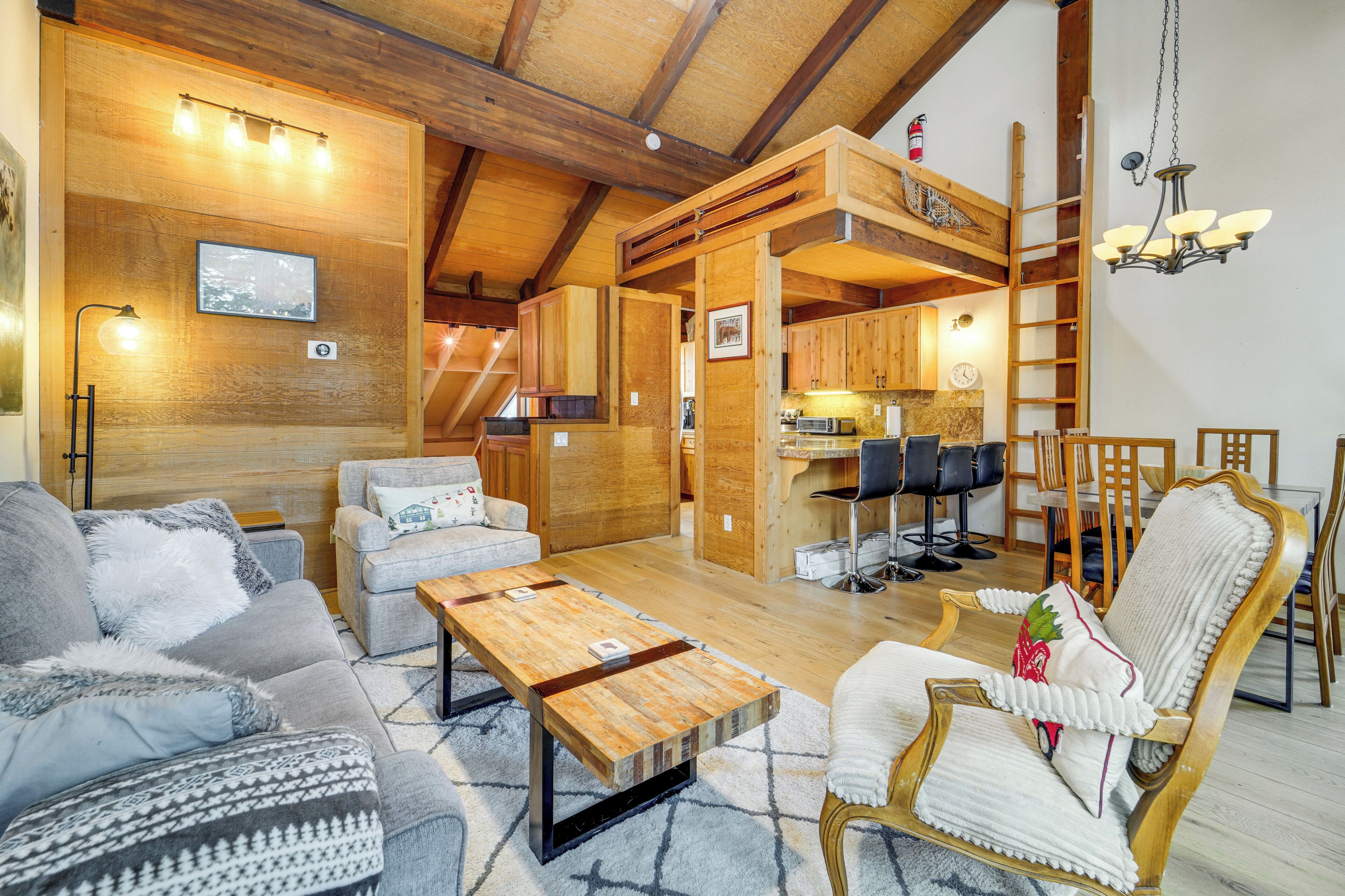 Un intérieur chaleureux avec des éléments en bois, comprenant un canapé, un fauteuil et une table basse. On aperçoit un coin cuisine au fond.