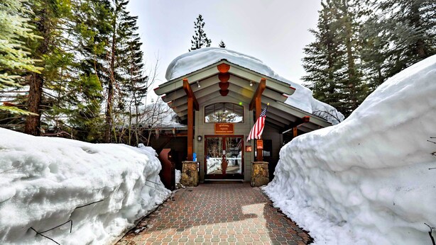 L’entrée d’un chalet, recouverte de neige, est bordée d’un chemin en briques menant à la porte. Des congères et de grands arbres l’entourent.