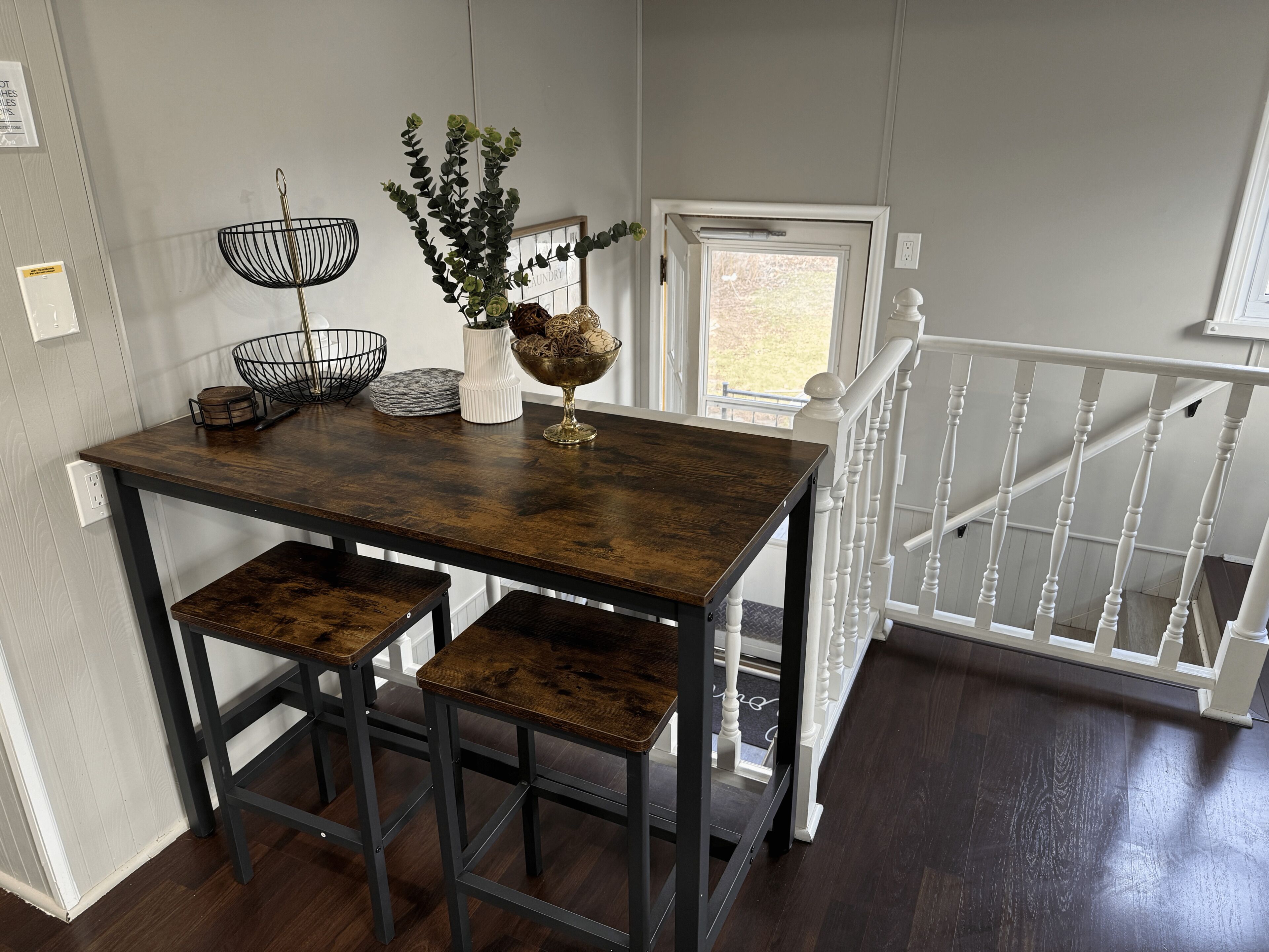 Un comedor con una mesa de madera oscura, dos taburetes a juego, un jarrón blanco con plantas verdes y una cesta de alambre negro.