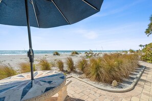 A beachside patio with a patterned table and a large umbrella, overlooking the ocean and beach. - Las Hermanas 1 Studio by RedAwning (Longboat Key)