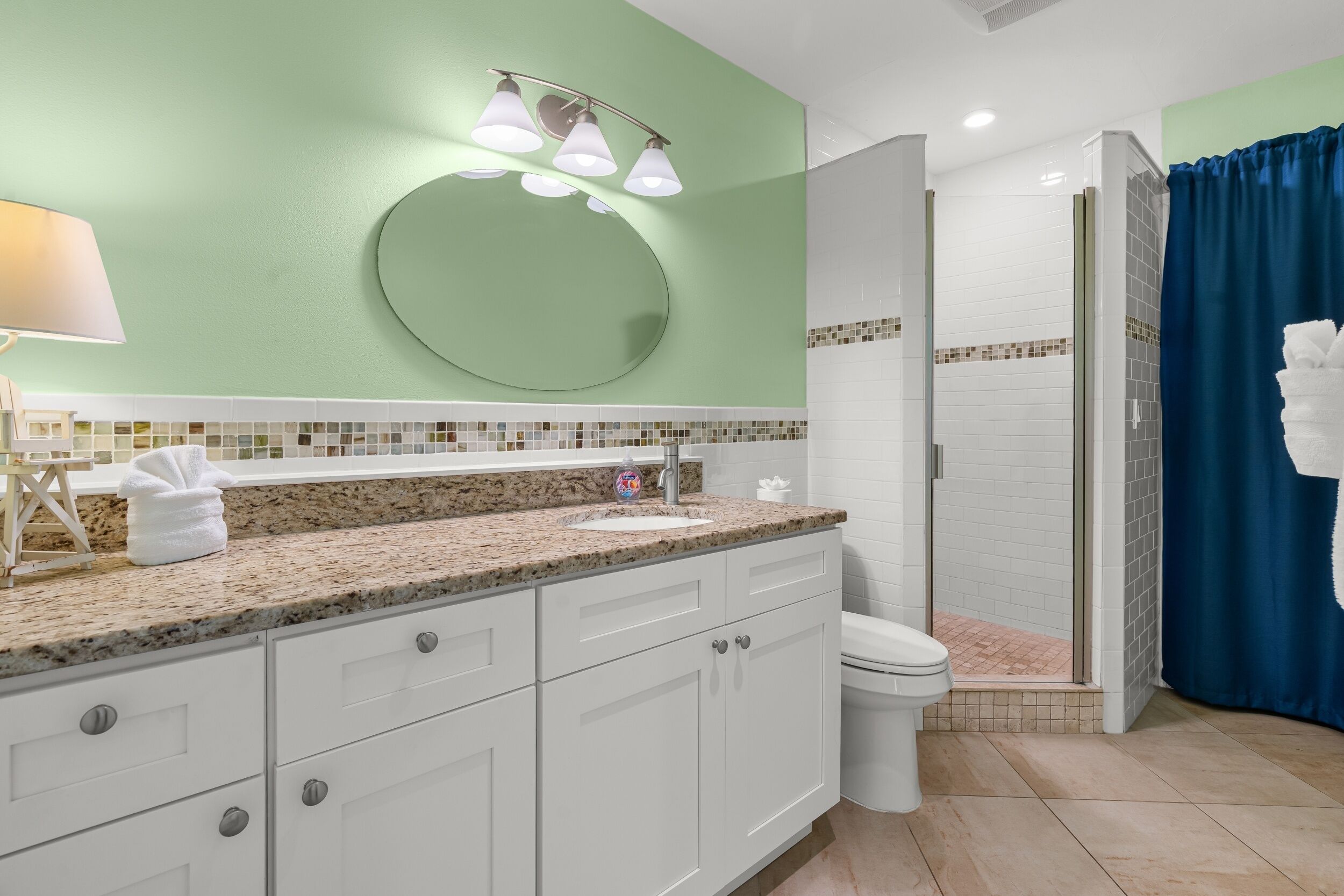 A bathroom with a granite countertop, white cabinets, a round mirror, and a shower with a blue curtain.