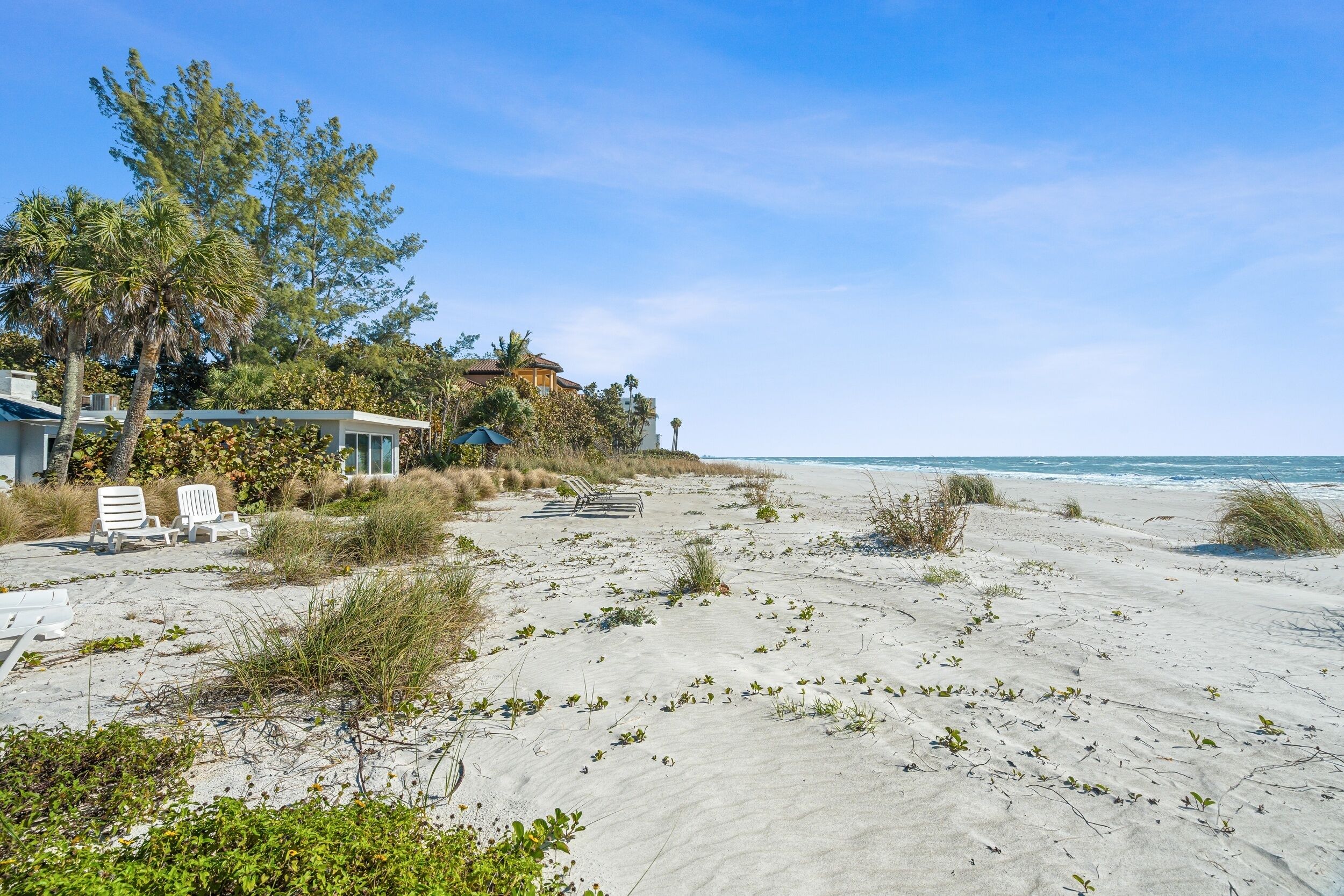 A beachfront property with a sandy beach, a few lounge chairs, and a house with a view of the ocean.