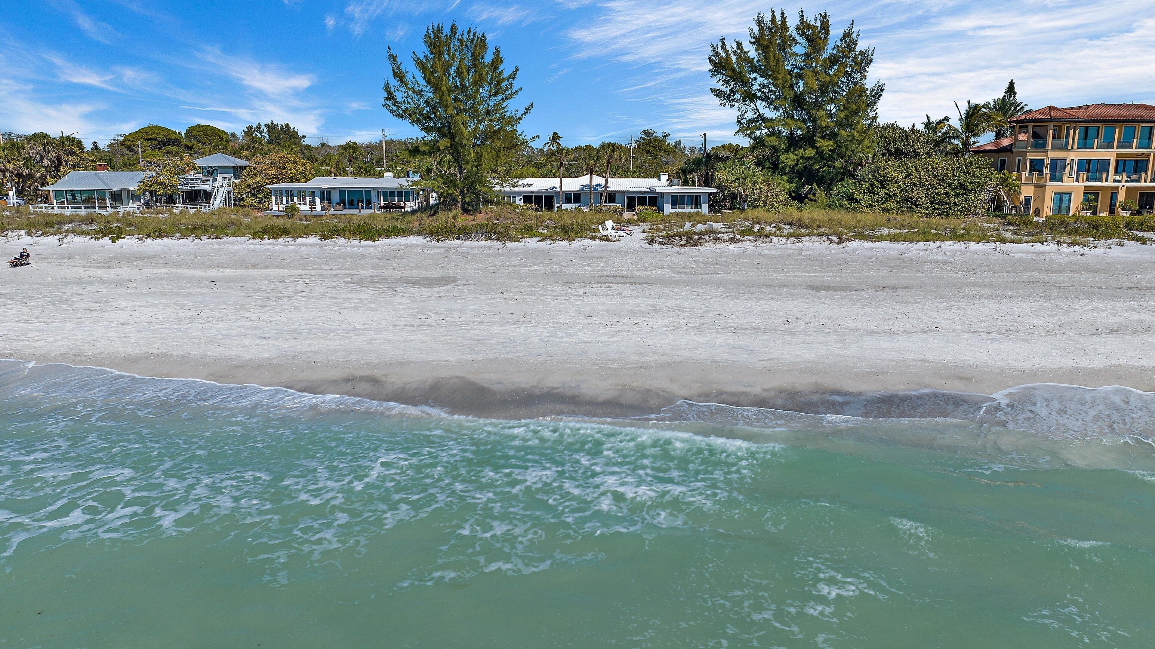 Une propriété située en bord de mer, avec une plage de sable, un ciel bleu dégagé et quelques bâtiments en arrière-plan.