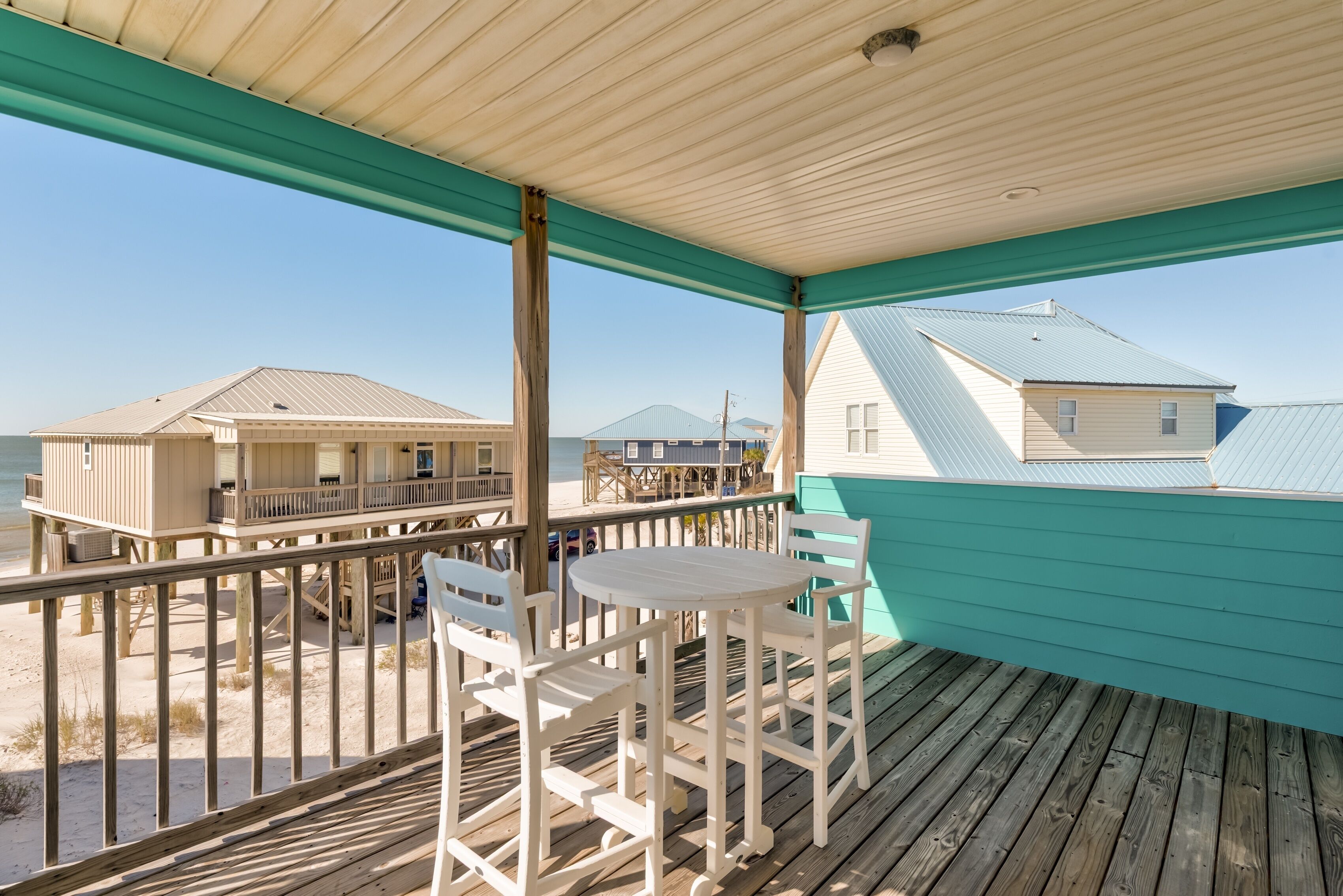 Eine Holzterrasse mit Tisch und Stühlen, mit Blick auf einen Strand und Gebäude im Hintergrund.