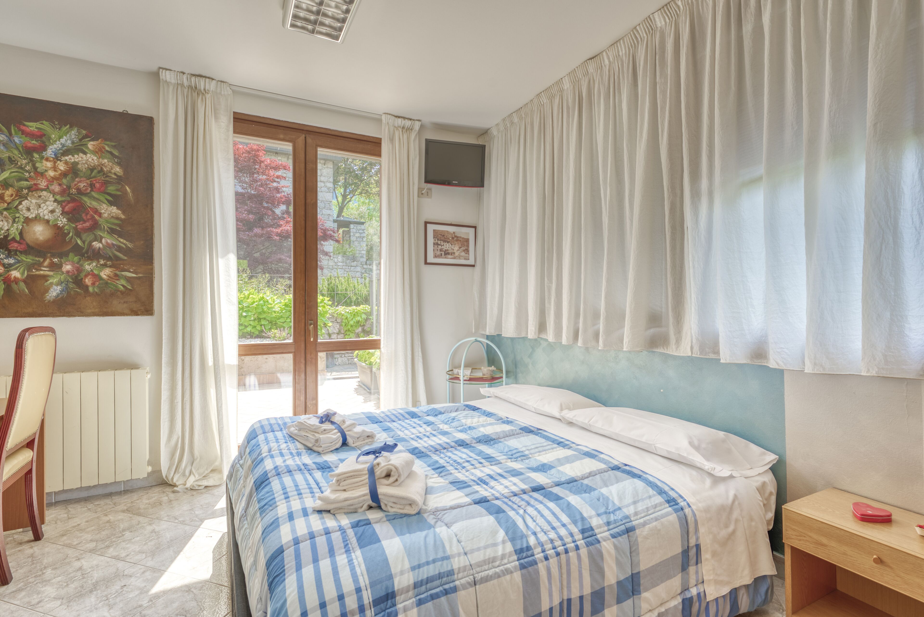 Une chambre avec une couvre-lit à carreaux, une table de chevet en bois, une chaise, un tableau au mur et une fenêtre avec des rideaux.