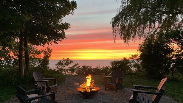 Ein Kiesbereich mit Feuerstelle und Adirondack-Stühlen, umgeben von Bäumen und mit Blick auf einen Sonnenuntergang über dem Wasser.