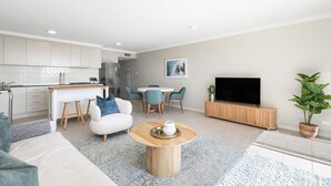 A modern living room with a sofa, coffee table, and TV. There's a dining area with chairs and a table. The kitchen has white cabinets and a tiled backsplash. - Panoramic views, 5-bedroom waterfront (Sylvania)